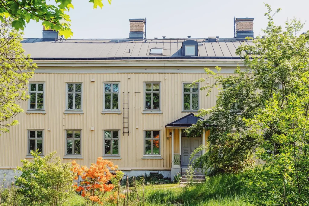 Gulmålad flerbostadsfastighet i trä med svart plåttak och trädgård i förgrunden, omgiven av grönska och blommande buskar.