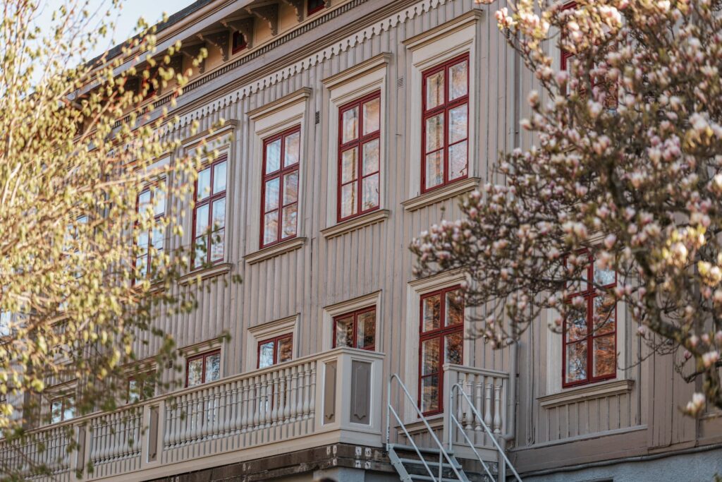 Träfasad med profilerade fönsteromfattningar och röda fönster, delvis skymd av blommande träd.